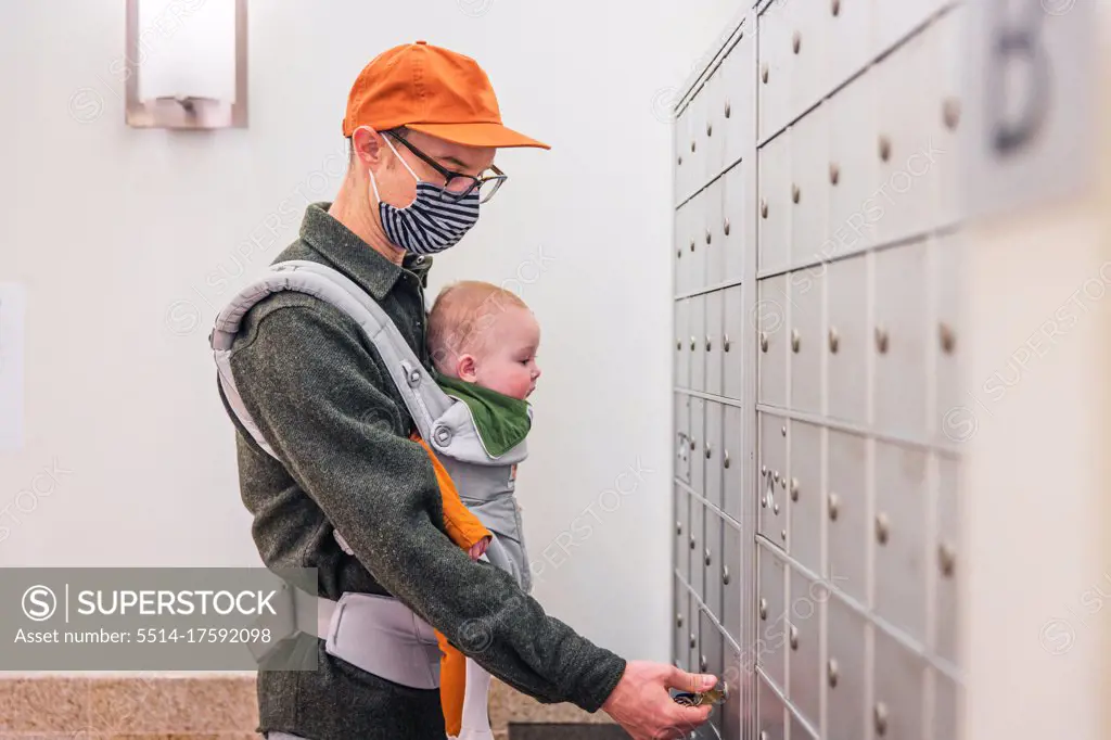 Father wearing face mask opening locker in room while carrying baby girl during COVID-19 crisis
