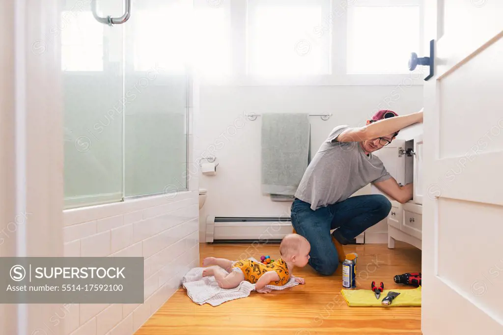Man fixing sink while daughter lying on hardwood floor in kitchen