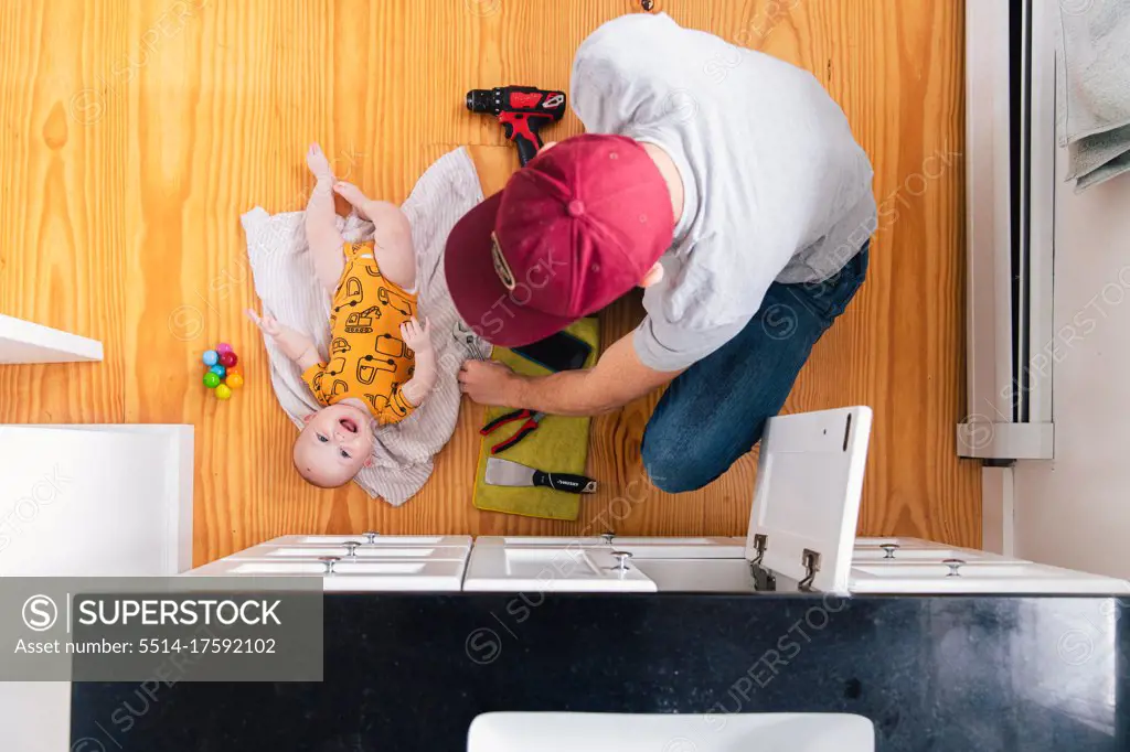 Overhead view of father fixing sink while happy baby girl lying on hardwood floor in kitchen at home