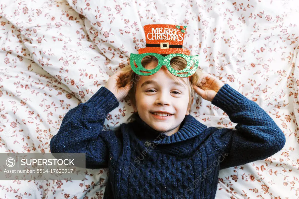 Little boy in a knit sweater with Christmas glasses lying on bed