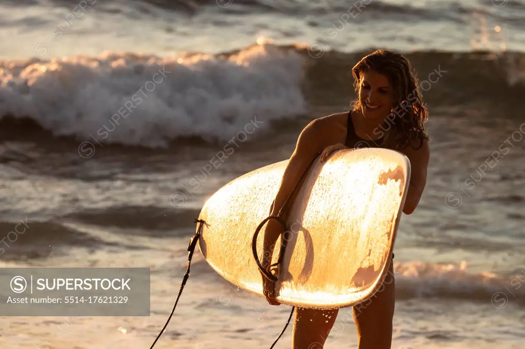 Woman carries surf board from the ocean at sunset in hawaii