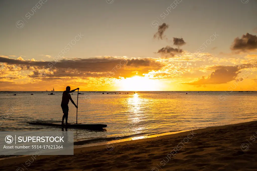 Male stand up paddle boarding drifts to the sand at sunset