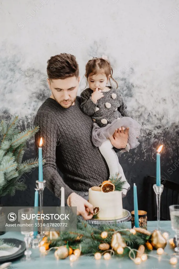 Young family with a daughter in festive outfits at a served table with candles, garlands, sparklers and a cake near the Christmas tree on New Years Eve