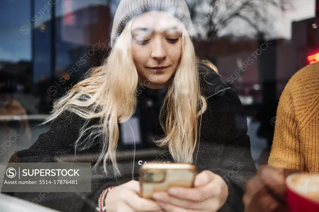 young female checking her phone, sitting at the window of a cafe with the city reflection on the glass. focus on the girl