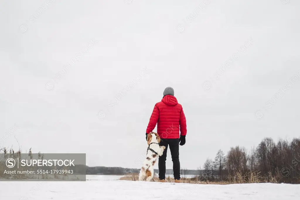 A dog stands next to his owner outdoors by a frozen snowy river.