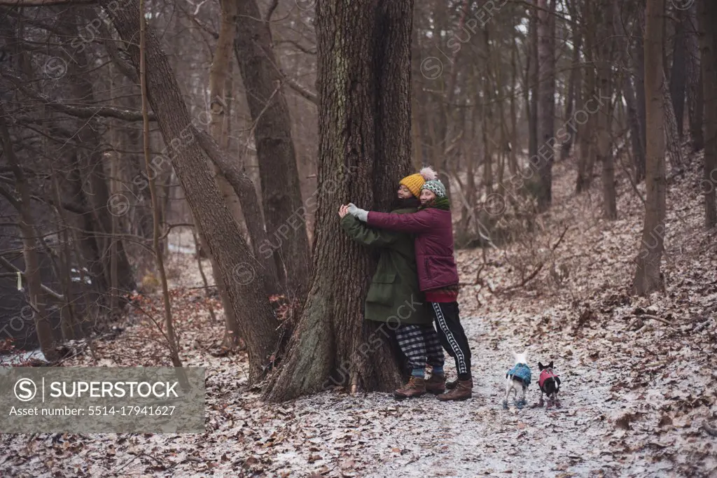 Lesbian couple hug a tree together with small dogs in winter germany
