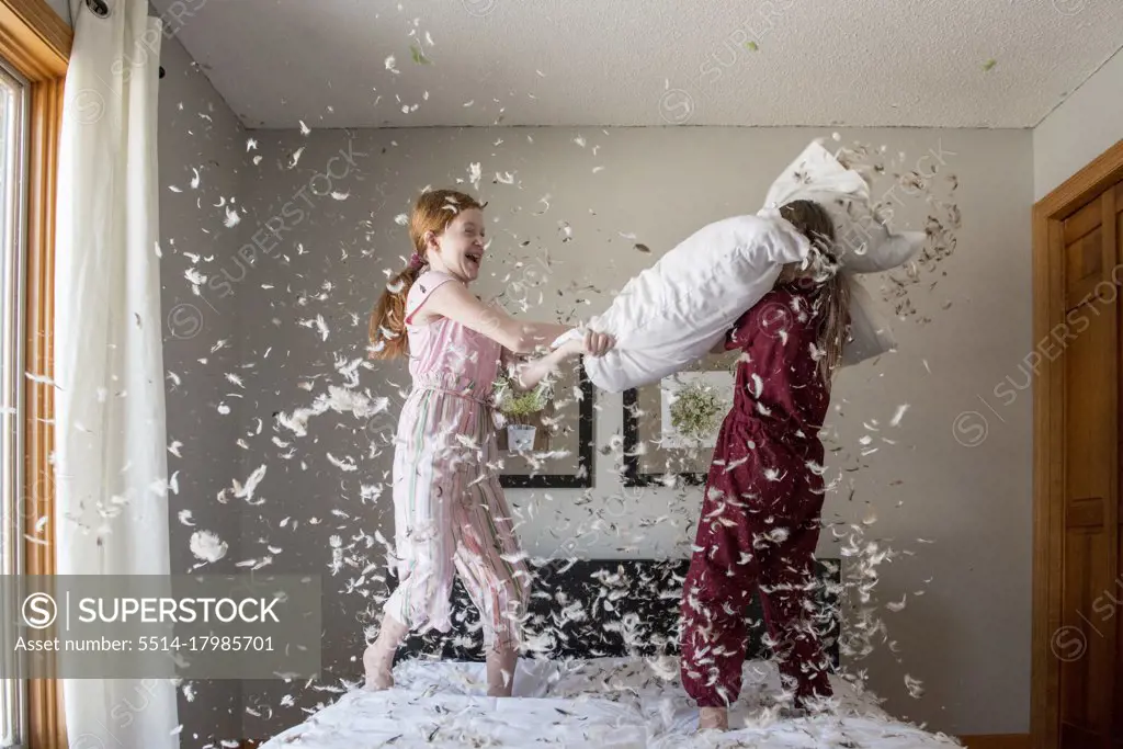 Two happy young girls having a feather pillow fight on the bed.