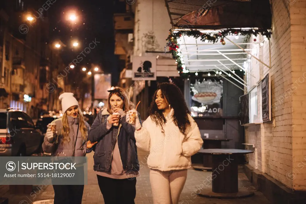 Female friends having hot drinks walking on street in city at night