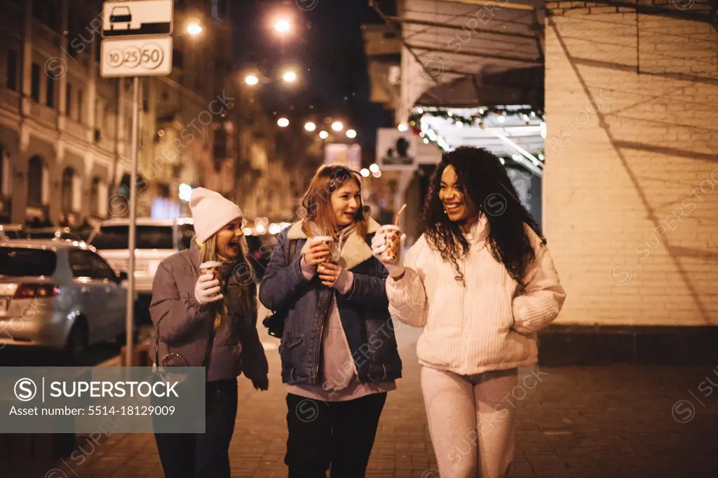Happy female friends having hot drinks walking on street in city