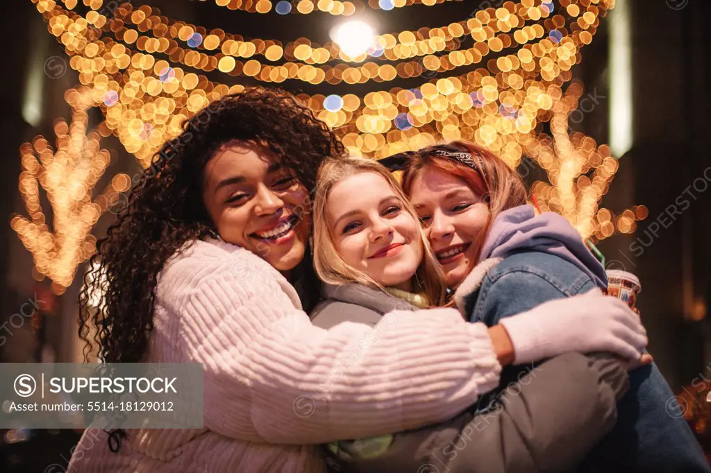 Happy female friends embracing standing by Christmas lights in city