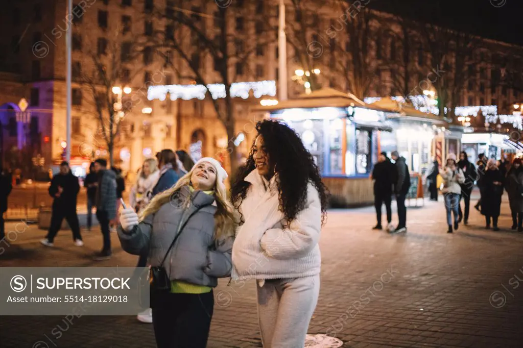 Happy female friends walking on street in city at night during winter