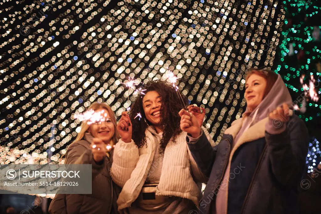 Happy female friends holding sparklers standing by Christmas lights