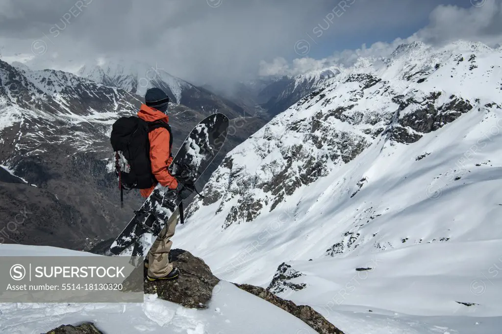 Man with snowboard standing on rock at snowcapped mountain against cloudy sky
