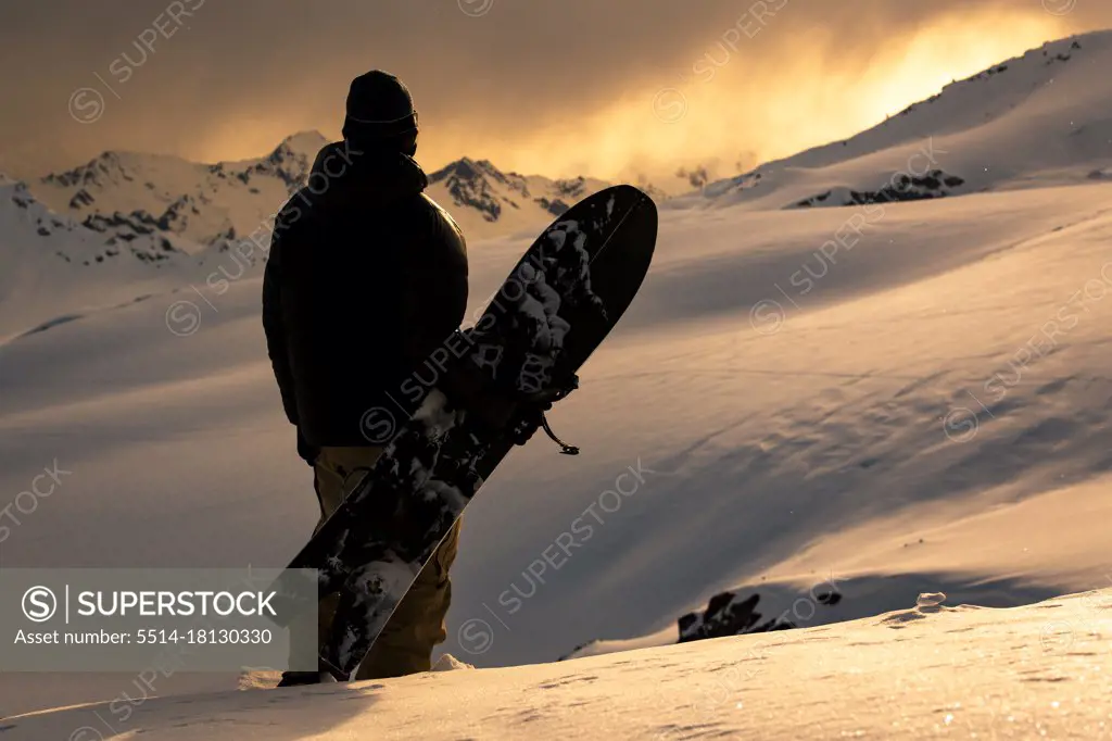 Rear view of man with snowboard standing on snowcapped mountain during sunset