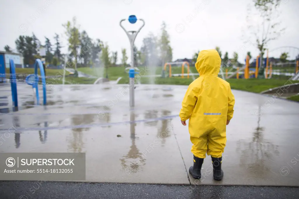 Toddler in rain suit and boots looks at the water park on a wet day