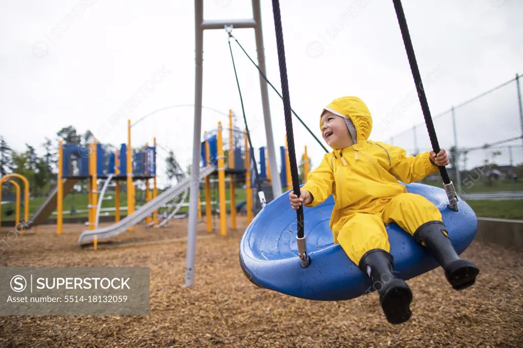 Happy toddler boy rides on swing at the park on a wet day