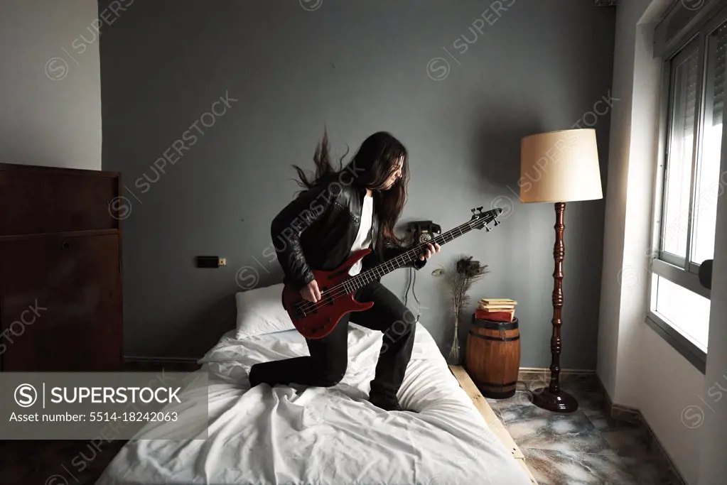 A man with long hair playing bass guitar on the bed