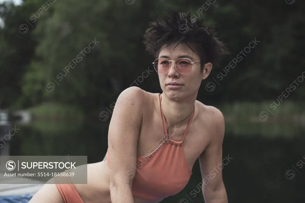 Athletic mixed-race woman in orange swimsuit sits on jetty over lake