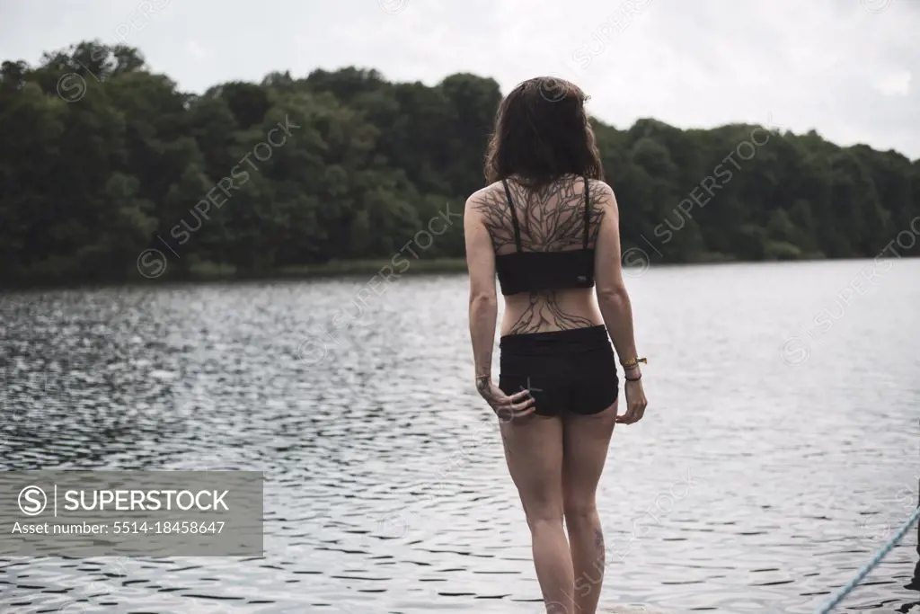 woman in with large tree tattoo stands at edge of natural lake