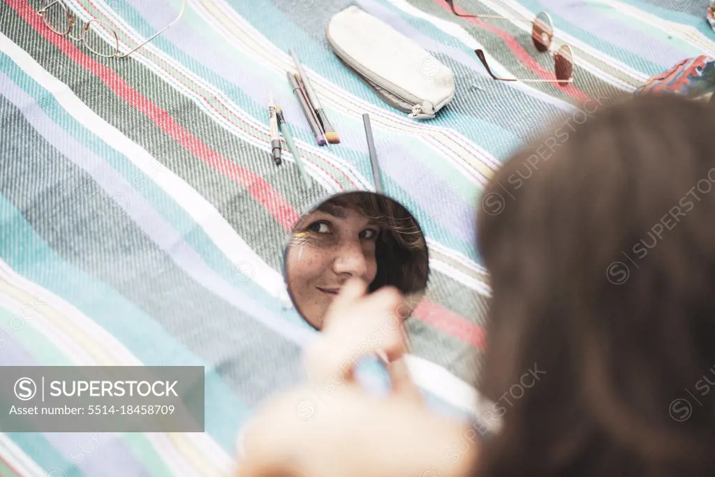 Artistic woman's reflection in mirror facepainting on summer blanket