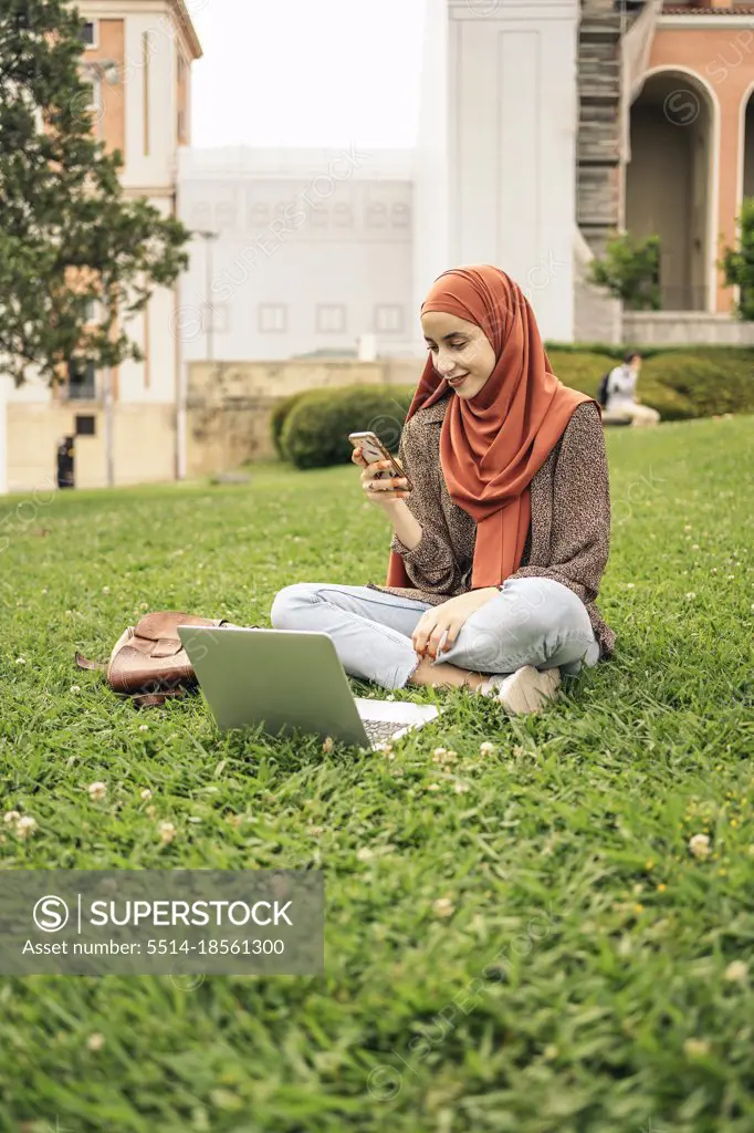 Muslim woman siting on the grass using a computer and phone