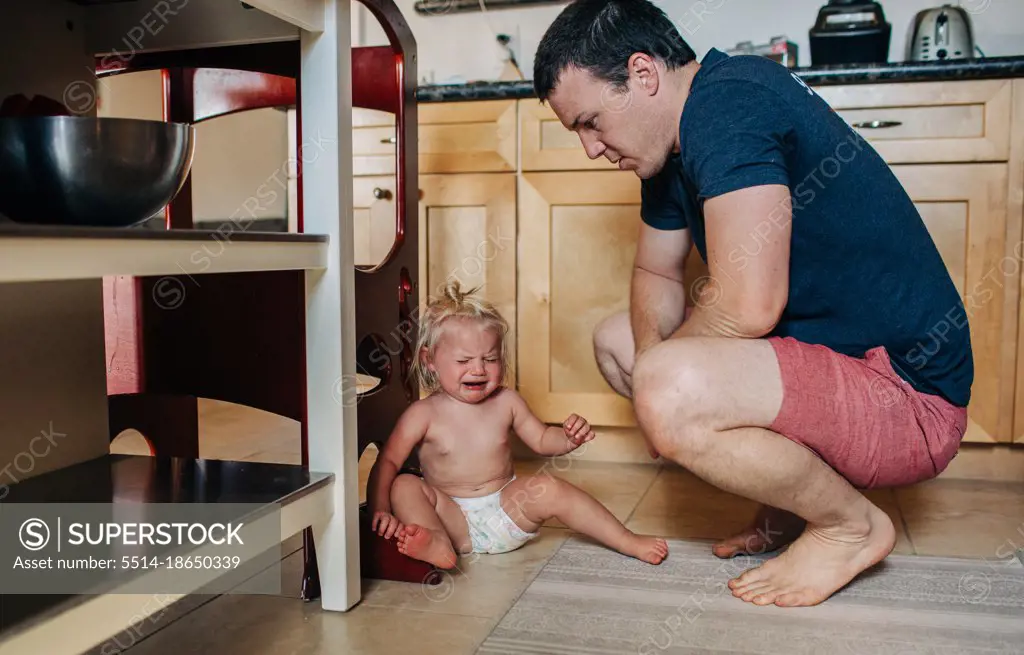 Dad looks down at diapered toddler girl while crying in kitchen