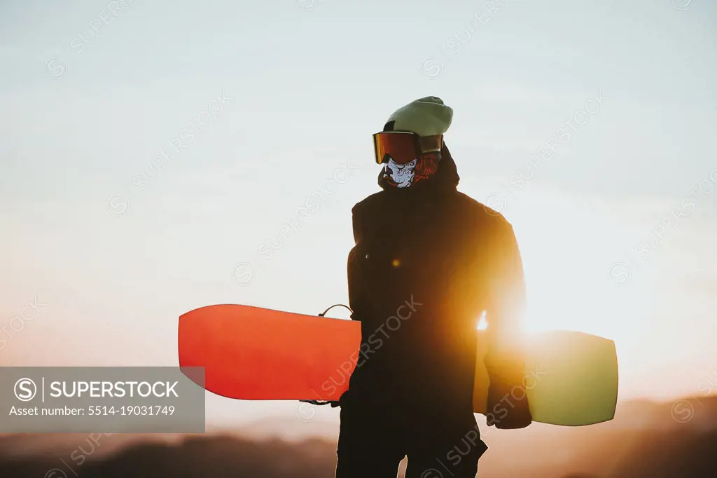Snowboarder in sportswear and glasses holding his snowboard at sunset