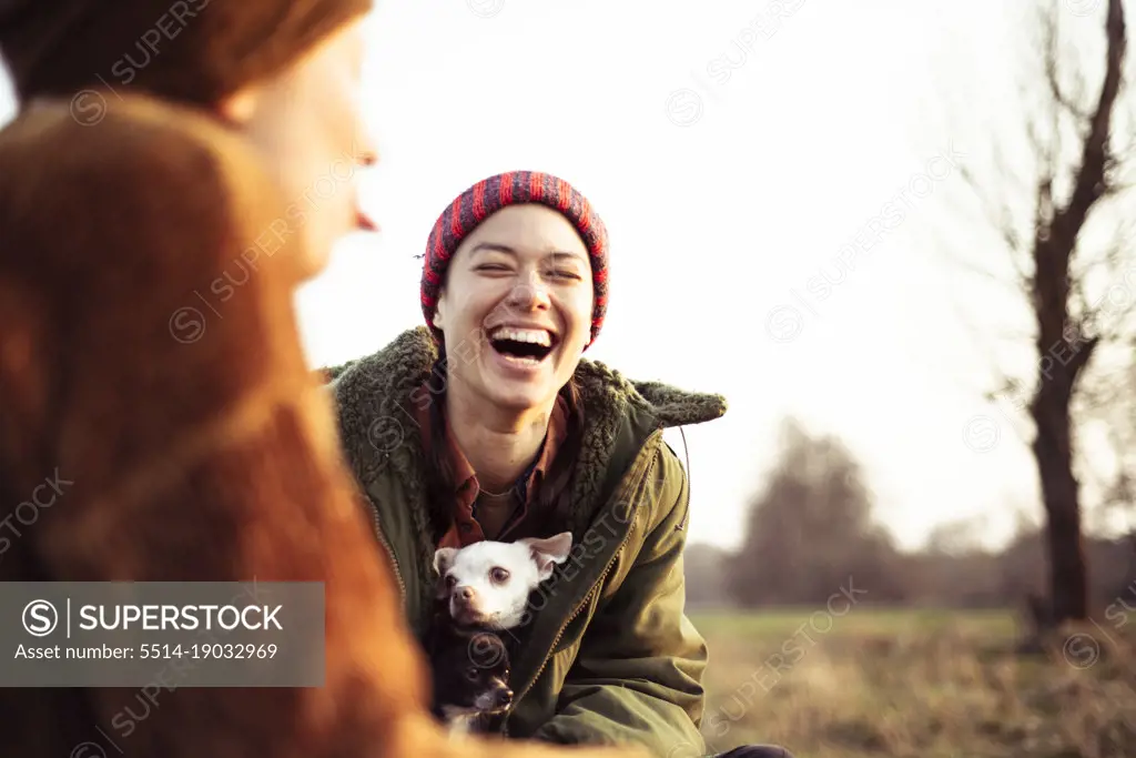 two friends laugh at joke with dogs in winter sun picnic in field