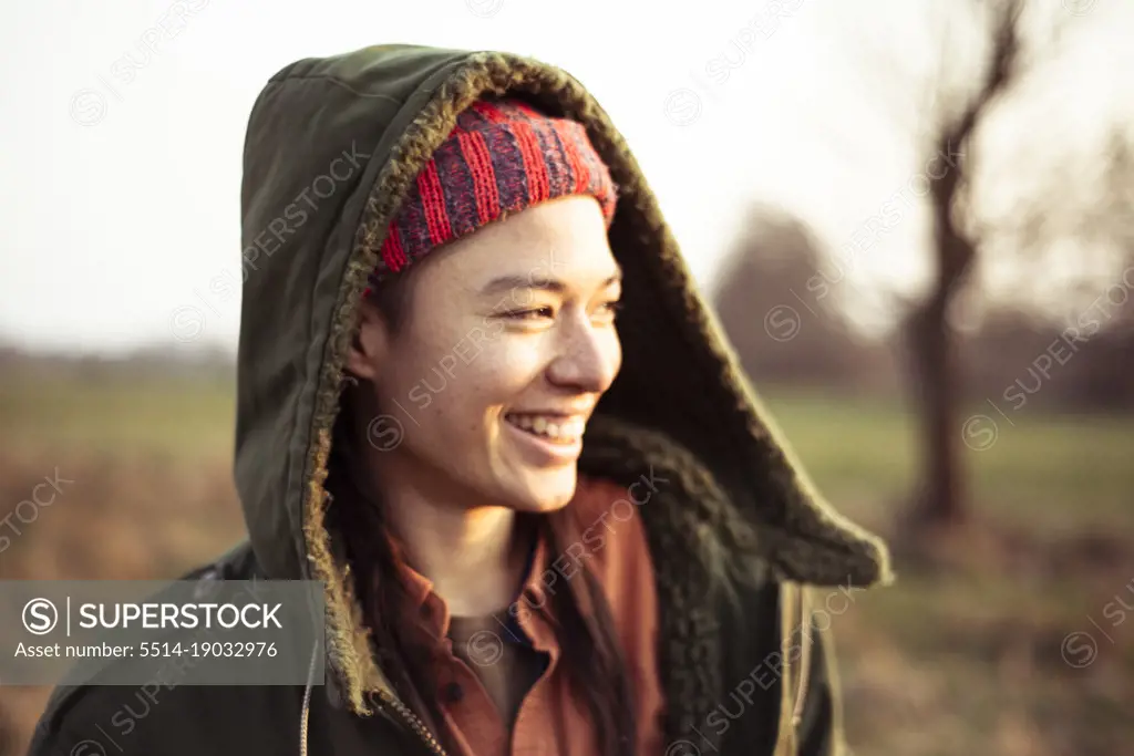non-binary person in warm jacket hood smiles in afternoon sun outdoors