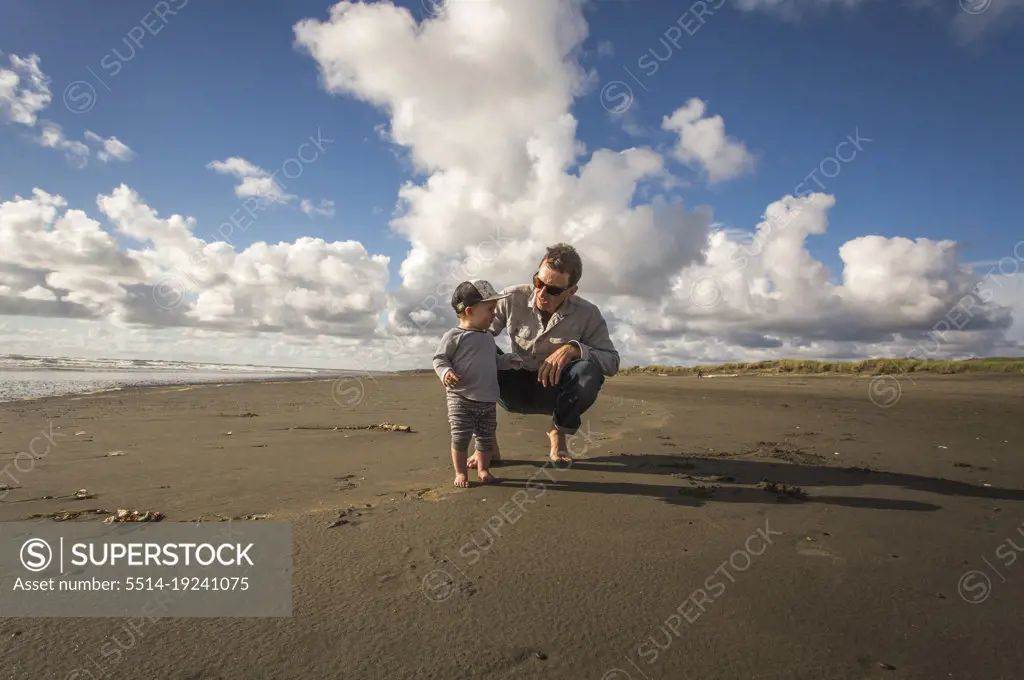 A man crouches to talk to a toddler on a wide empty beach