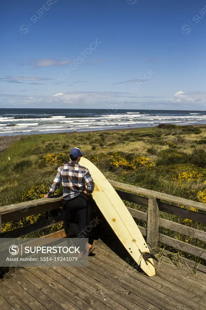 A man with a surfboard overlooks the beach from a deck