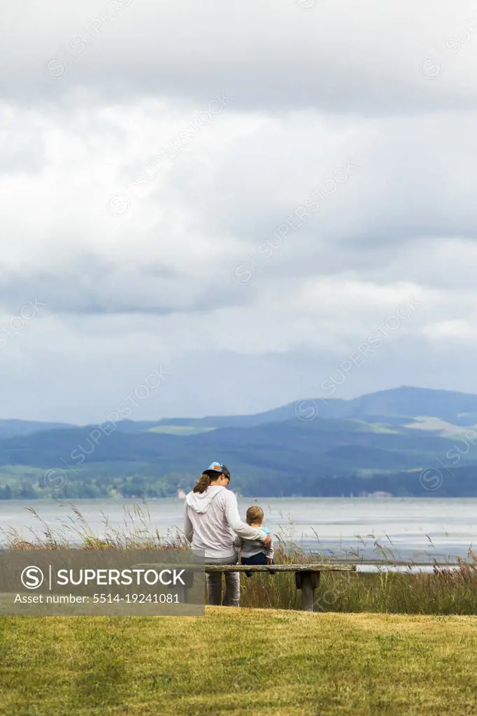 A woman sits beside a child on a bench overlooking water and mountains
