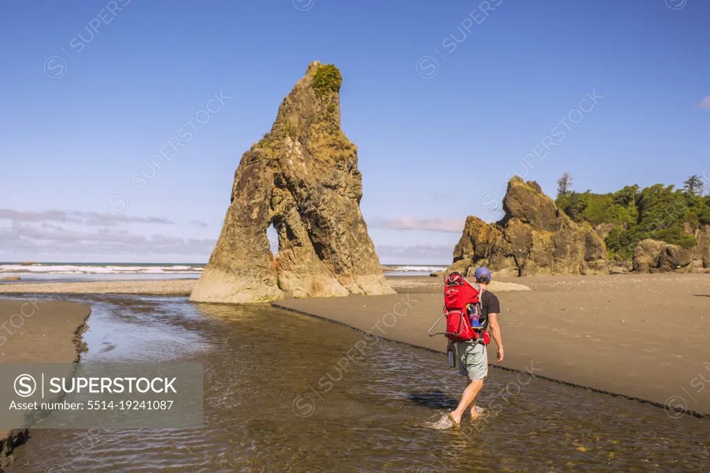A man walks across a stream on a beach carrying a toddler in a pack