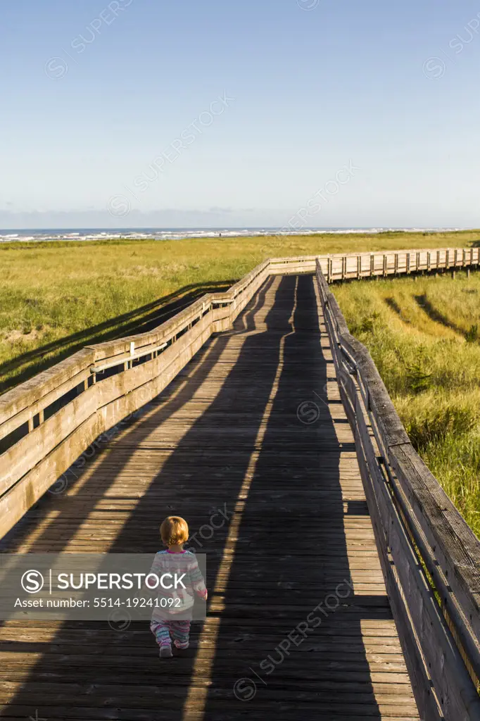 A small child walks down a long wooden boardwalk among grassy dunes