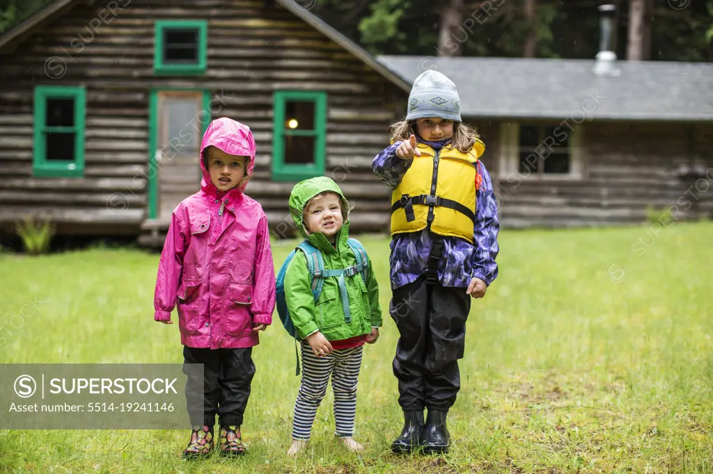 Three young kids in rain gear in front of a log cabin