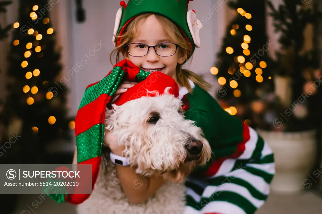 Girl hugs pet golden doodle dog in Santa hat on Christmas porch