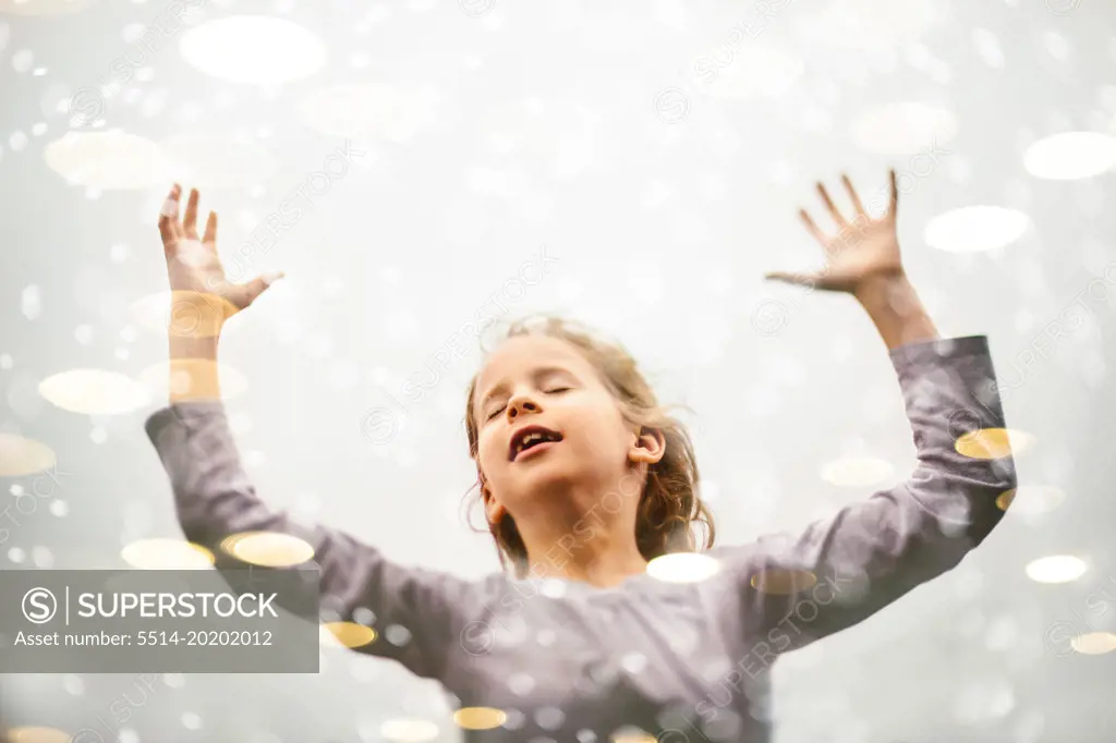 Blond girl child joyfully raises hands in air with light bokeh