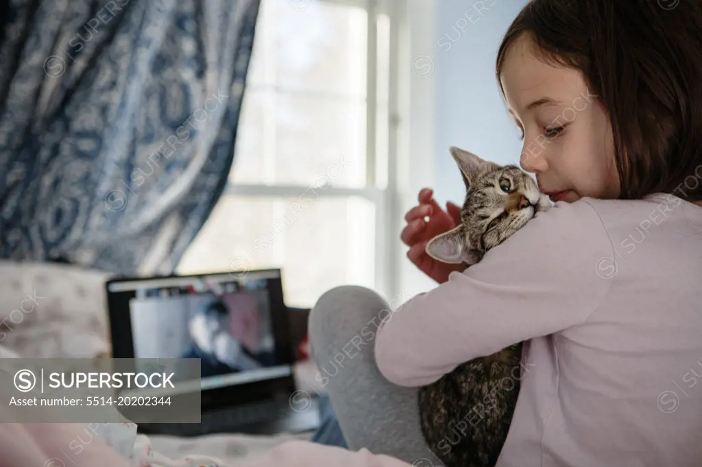 A little girl sits on bed in front of computer snuggling a kitten