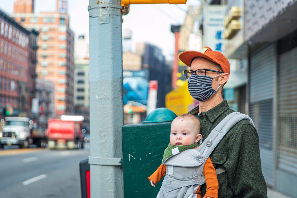 Father in face mask looking away while carrying baby girl on sidewalk in city during COVID-19 crisis