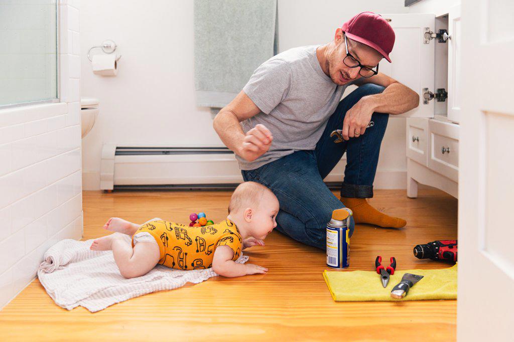 Father fixing kitchen sink while baby girl lying on hardwood floor at home