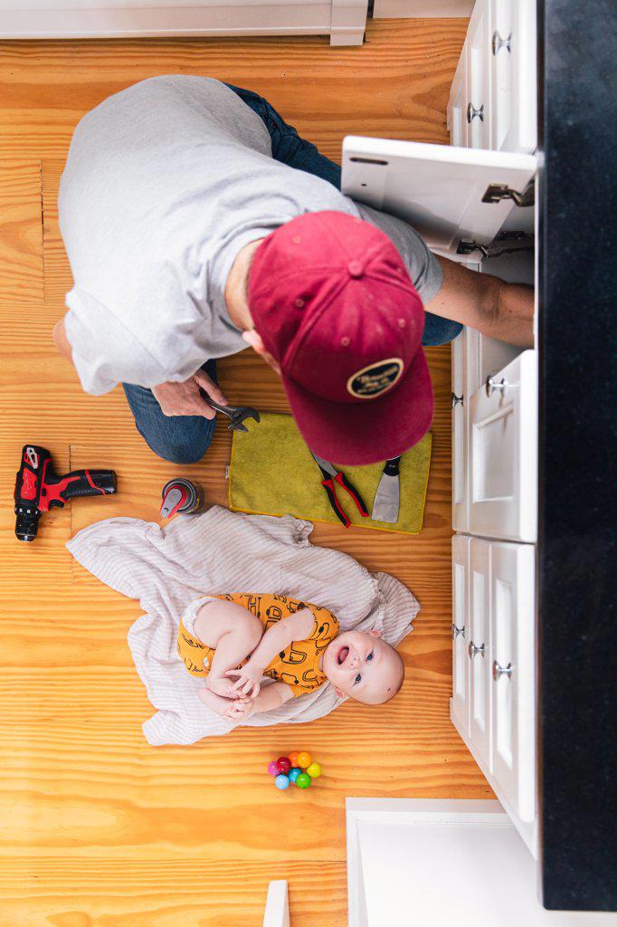 Overhead view of father fixing kitchen sink while baby girl lying on hardwood floor at home