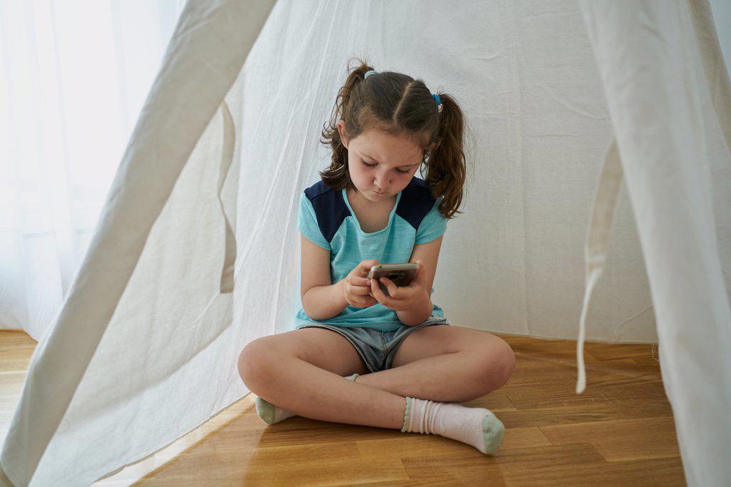 Little girl looking at her smartphone inside a white teepee tent inside her house. Technology concept