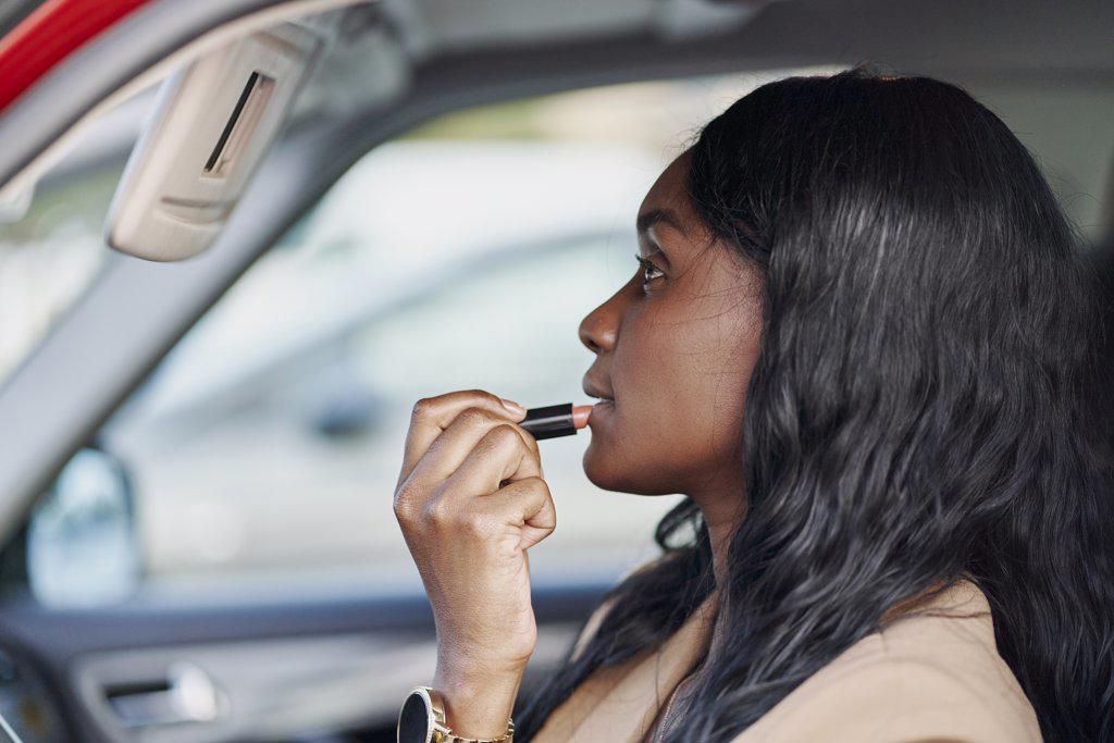 Portrait of a black woman in a brown suit painting her lips inside her car. Business concept and elegance