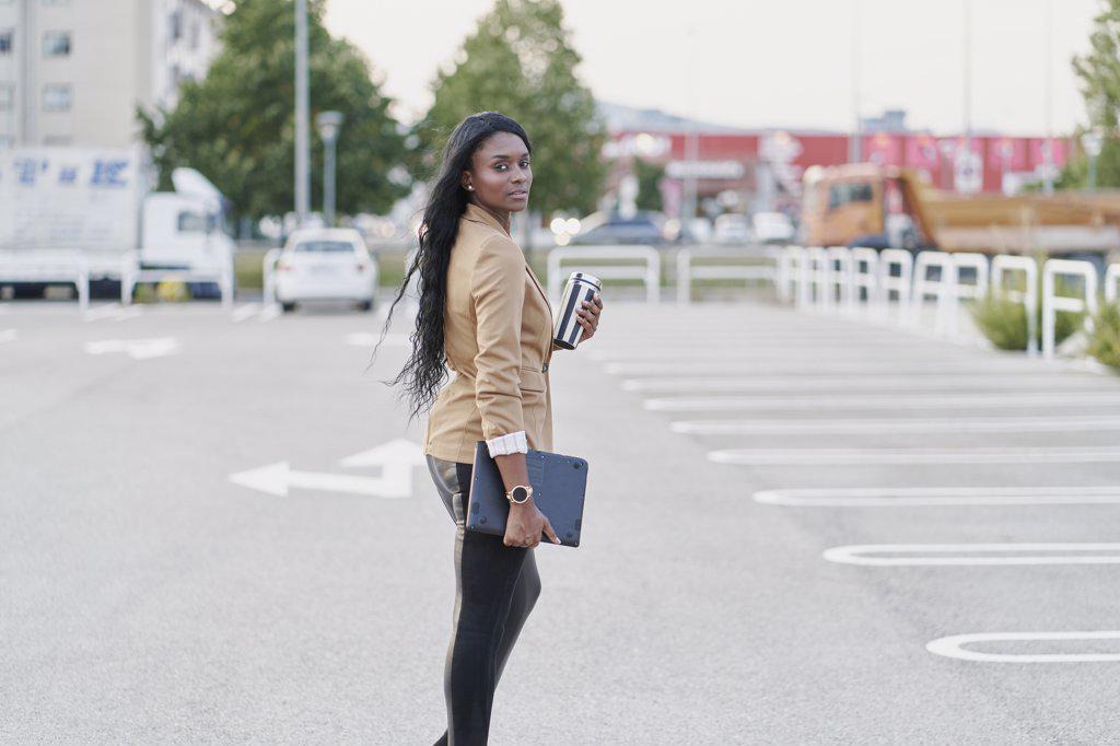 Portrait of black woman wearing a brown suit holding a laptop and a container with coffee walking down the street