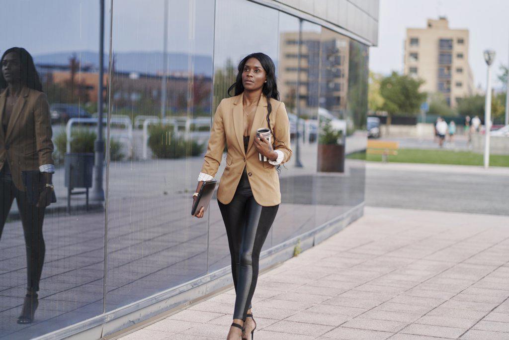 Black woman wearing a brown suit holding a laptop and a container of coffee walking next to a building. business concept