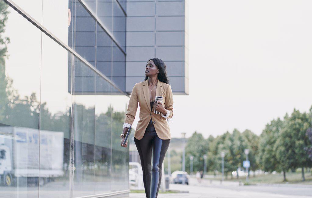 Black woman wearing a brown suit holding a laptop and a container of coffee walking next to a building. business concept