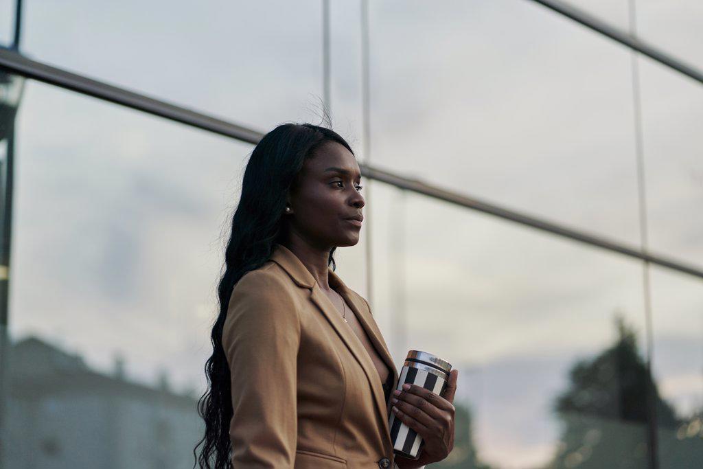 side view of a black woman wearing a brown suit holding a laptop and a container of coffee walking next to a building. business concept