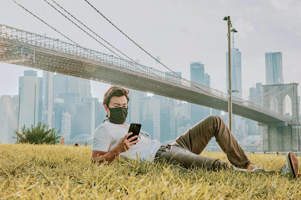 Young man sitting in park wearing face mask on phone.