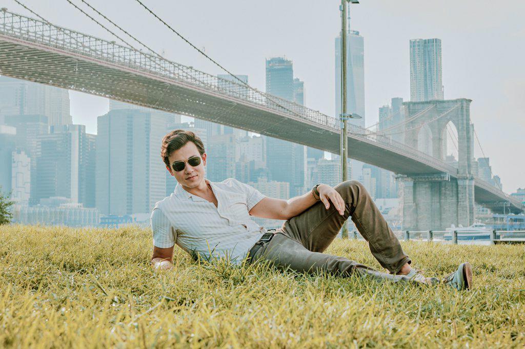 Young man sitting outdoors in park wearing sunglasses.