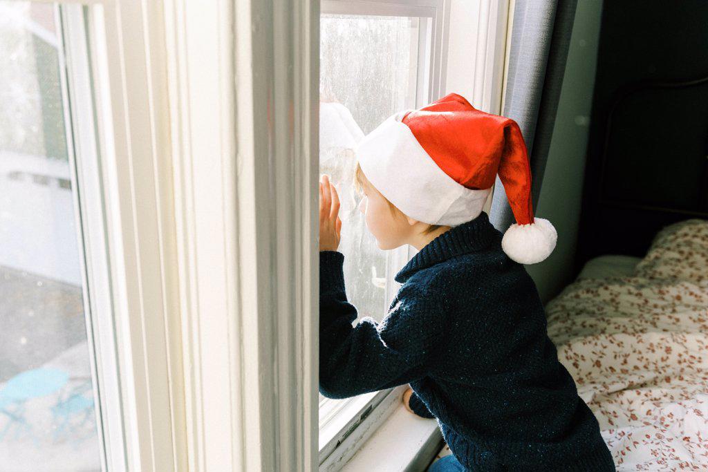 Little boy with Christmas hat looking out the window for Santa clause
